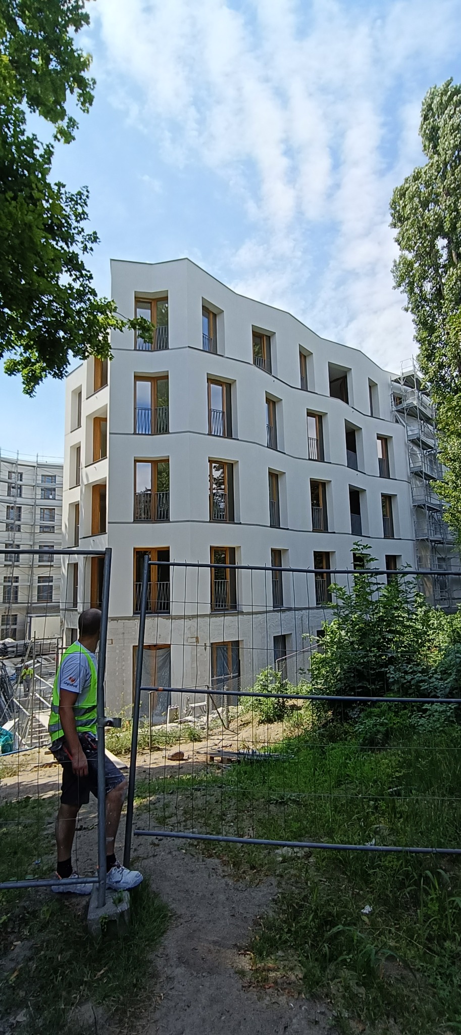 Wielopiętrowy budynek w trakcie prac wykończeniowych elewacji, widoczne rusztowania i pracownik w kamizelce odblaskowej przy ogrodzeniu placu budowy.