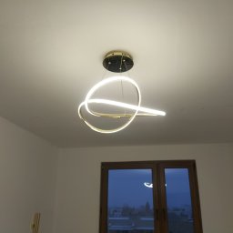 STANINSTAL-TECH - Nowoczesna lampa sufitowa LED o zakrzywionym kształcie, zainstalowana w pokoju z widokiem na miasto przez okno.