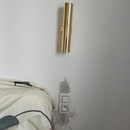 STANINSTAL-TECH - Złota, cylindryczna lampa ścienna nad gniazdkiem elektrycznym z widocznymi śladami po malowaniu na ścianie, obok fragmentu łóżka z wzorzystą pościelą.