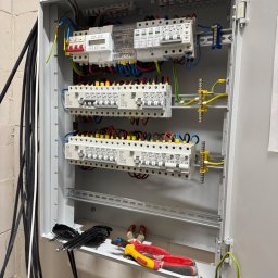 STANINSTAL-TECH Stanislav Udodov - Rozdzielnica elektryczna z zamontowanymi bezpiecznikami, przewodami i licznikiem. Widoczne narzędzia. Instalacja w szarej obudowie na tle ściany z betonu komórkowego.