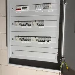 Szara, otwarta skrzynka elektryczna z bezpiecznikami i oznaczeniami, zamontowana na murze z białych bloczków. Widoczne kable wewnątrz.