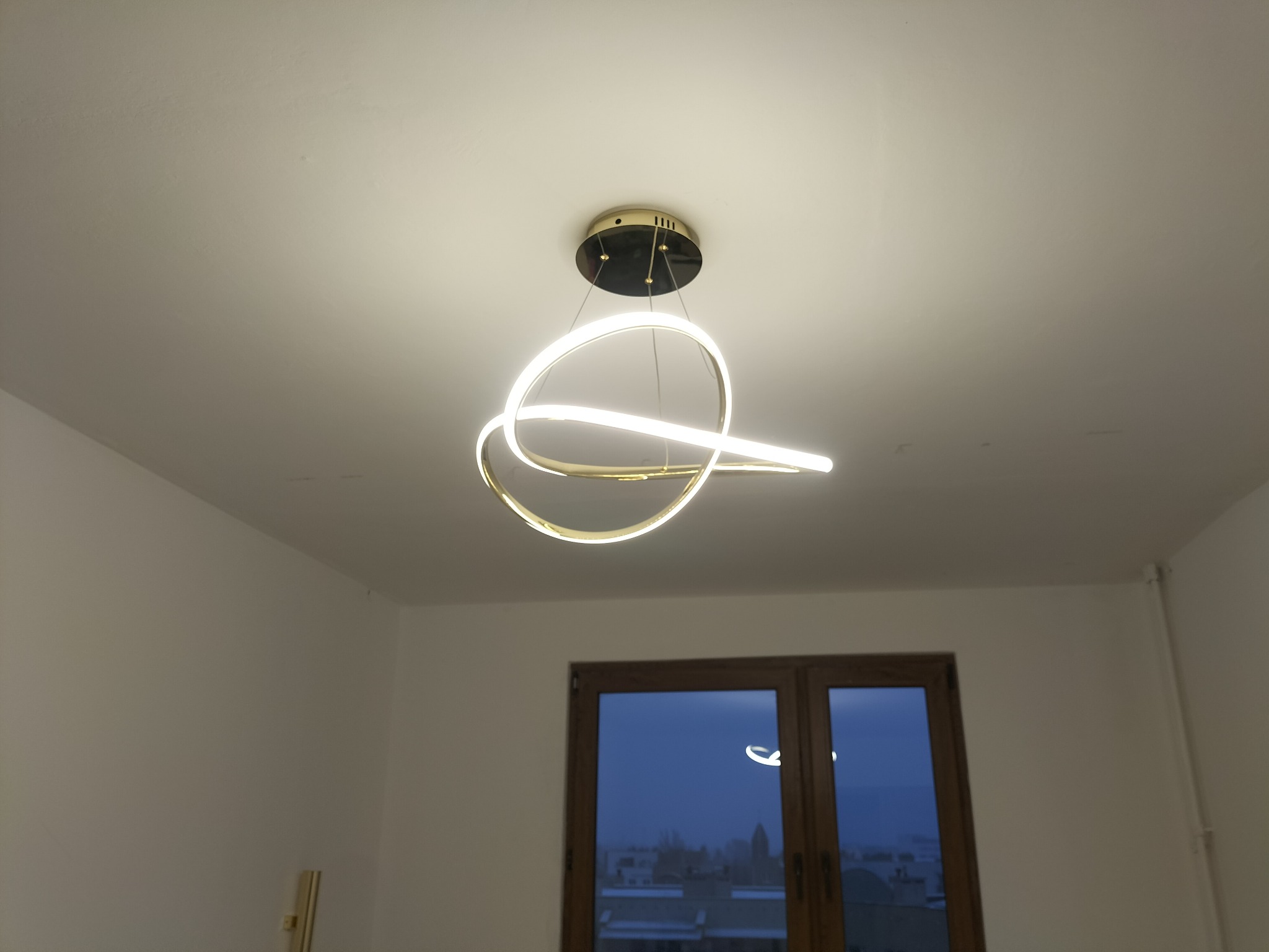 Nowoczesna lampa sufitowa LED o zakrzywionym kształcie, zainstalowana w pokoju z widokiem na miasto przez okno.