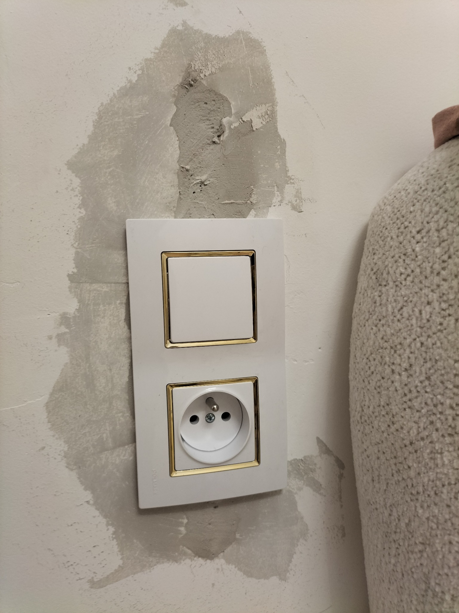 Biały włącznik światła i gniazdko z złotymi ramkami zamontowane na ścianie z widocznymi śladami po szpachlowaniu.