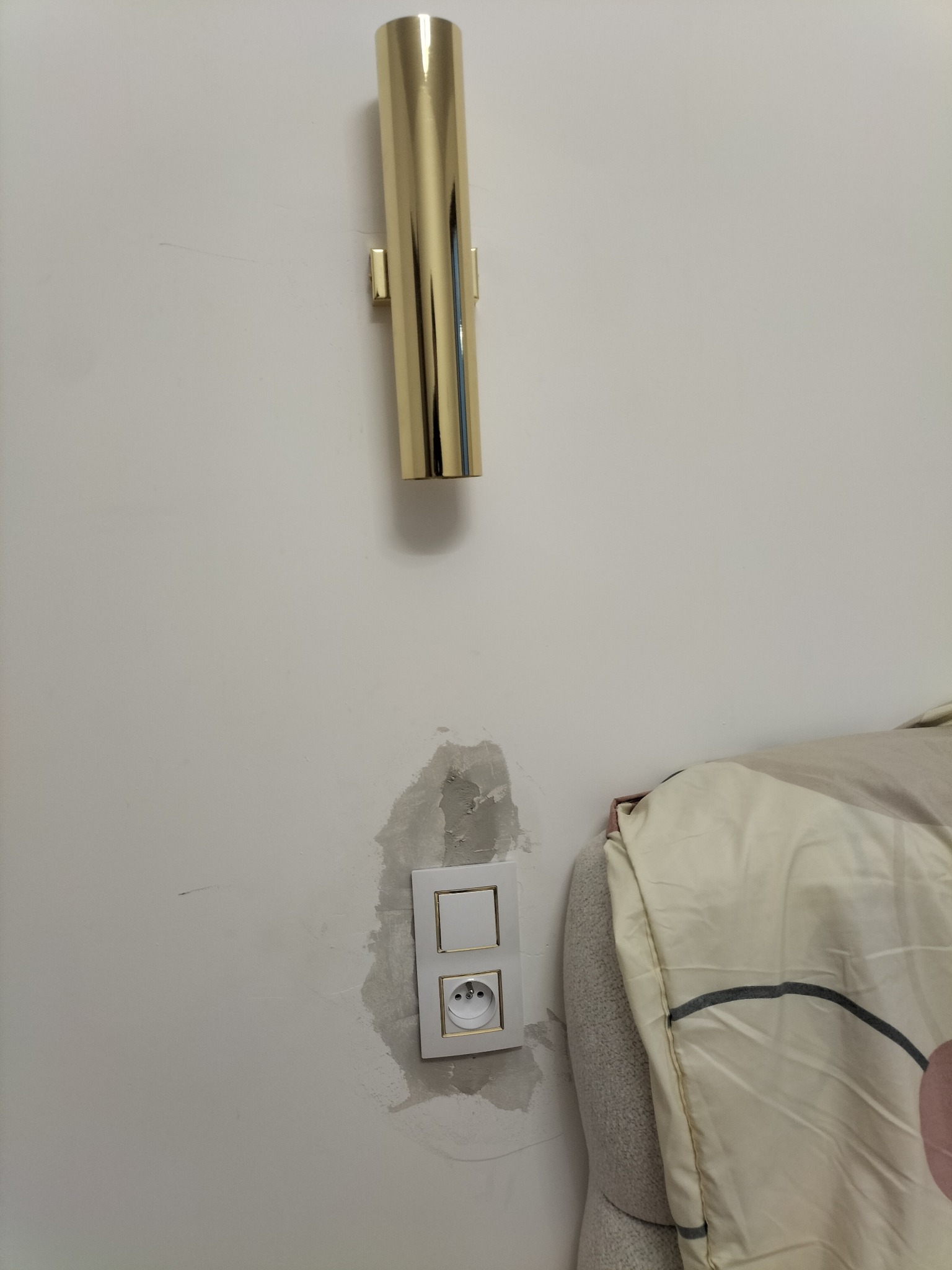 Ściana z zamontowanym złotym kinkietem tubowym i gniazdkiem elektrycznym z widocznymi śladami po montażu, obok fragment łóżka z kołdrą.