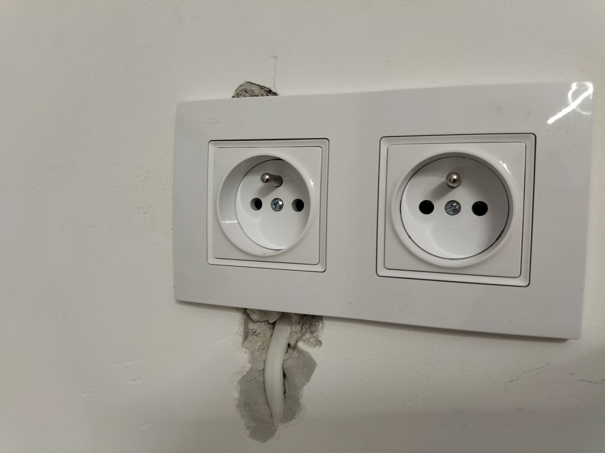 Podwójne, białe gniazdko elektryczne typu E zamontowane w ścianie, widoczne niedoskonałości w tynku wokół kabla.