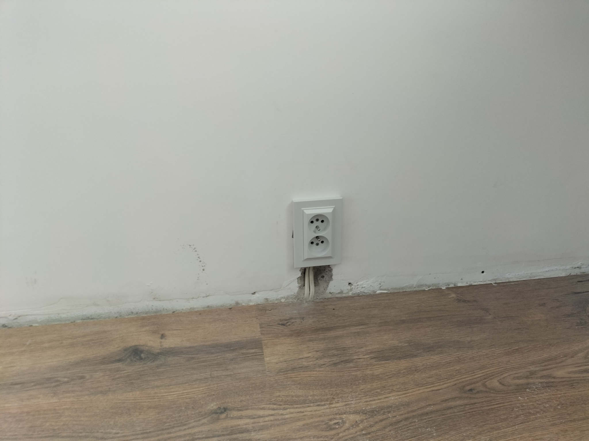 Białe gniazdko elektryczne z dwoma otworami zamontowane w ścianie nad brązową podłogą, widoczne przewody w ścianie, brak listwy przypodłogowej, ślady po remoncie na ścianie.