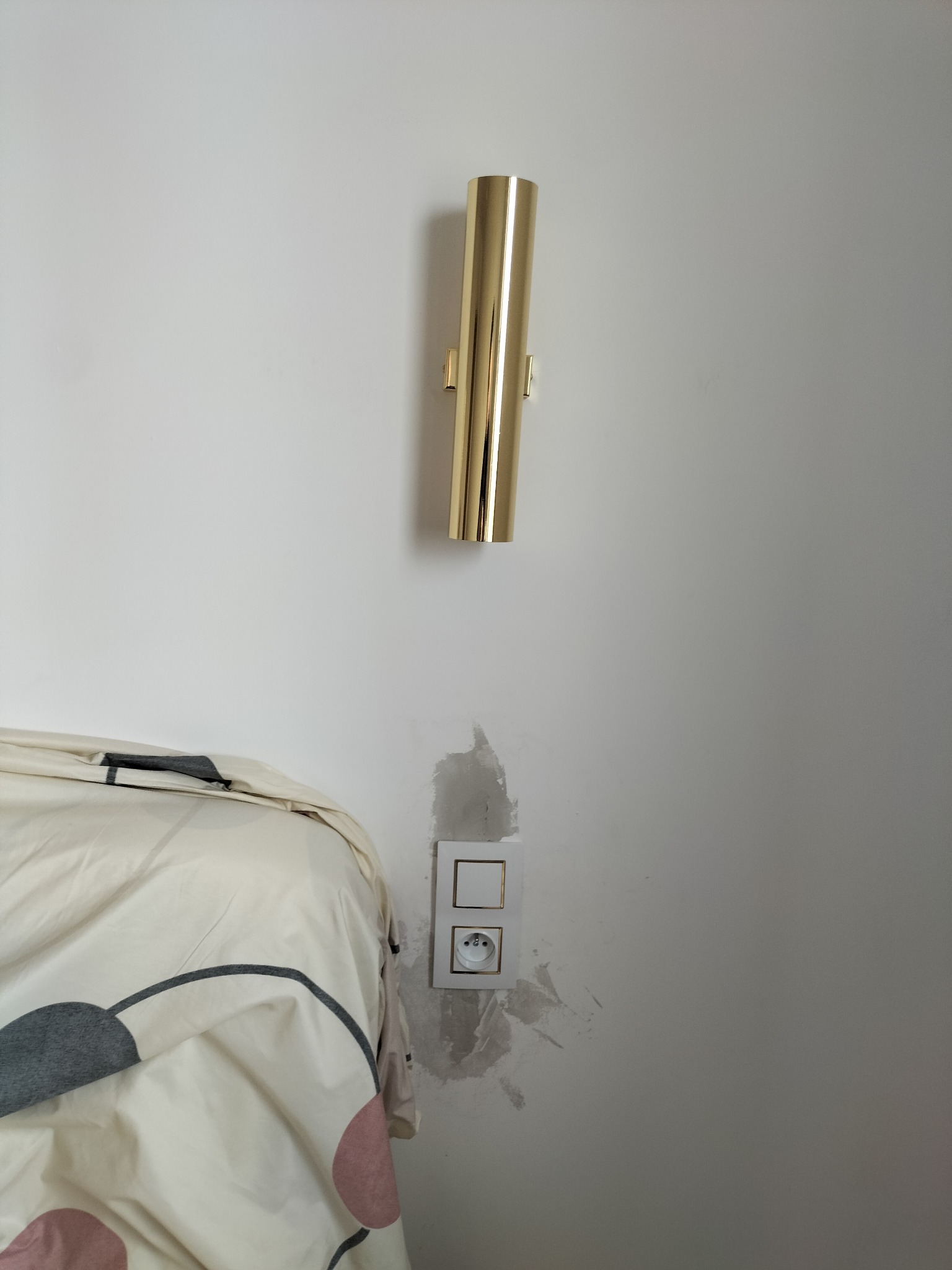 Złota, cylindryczna lampa ścienna nad gniazdkiem elektrycznym z widocznymi śladami po malowaniu na ścianie, obok fragmentu łóżka z wzorzystą pościelą.
