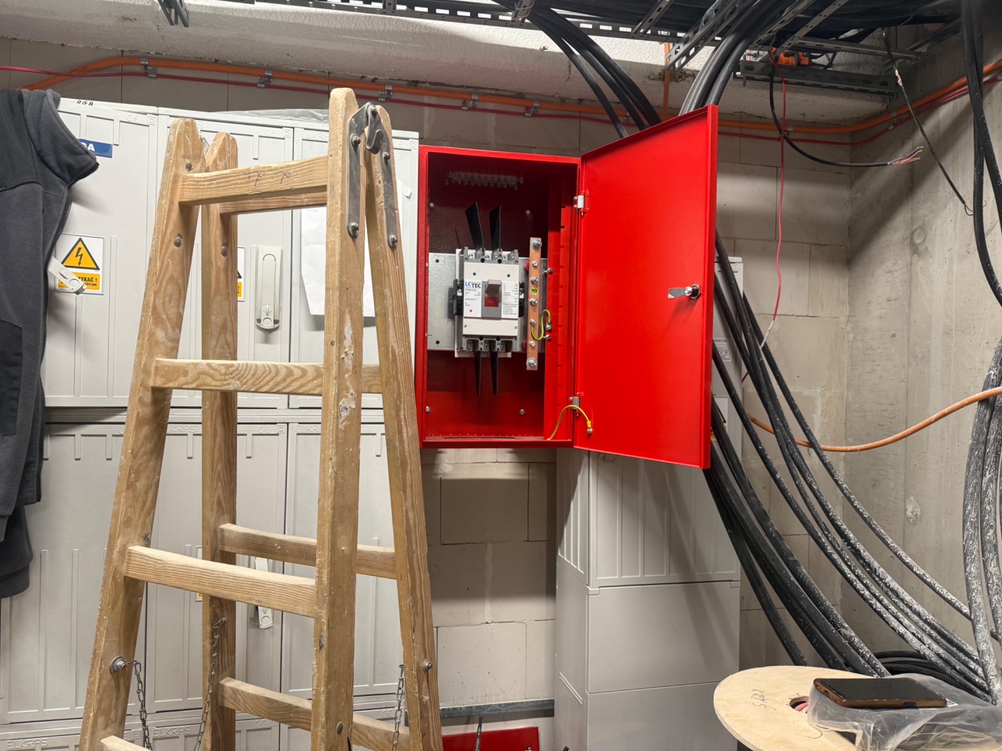 Otwarta, jaskrawoczerwona szafka elektryczna z widocznym wyłącznikiem, zamontowana na ścianie z bloczków. Obok stoi drewniana drabina, a nad nią widoczne kable.
