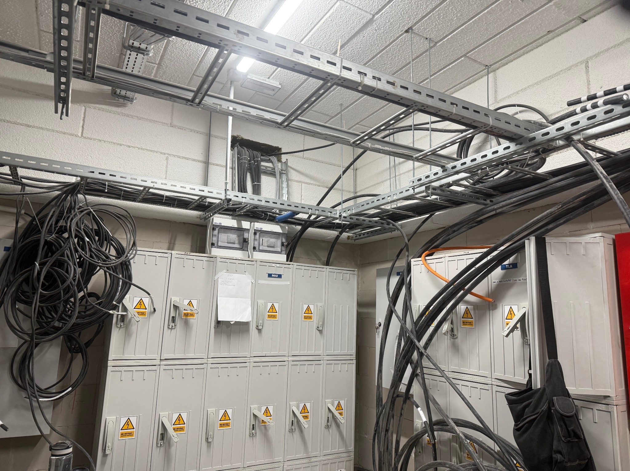 Rozbudowana instalacja elektryczna z szynami montażowymi pod sufitem i szafami rozdzielczymi z ostrzeżeniami. Widoczne przewody i elementy zabezpieczeń.