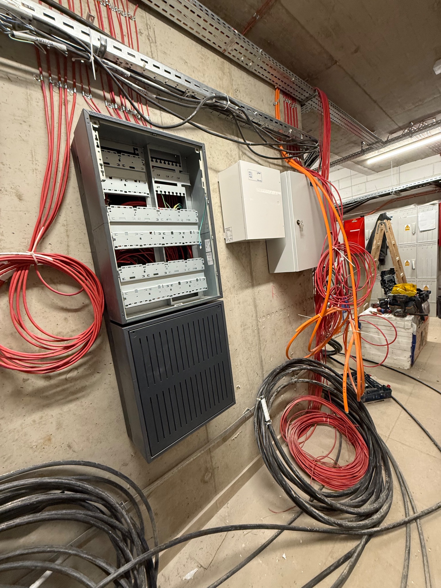 Instalacja elektryczna w budynku w trakcie montażu: szafa rozdzielcza z podłączonymi kablami różnych kolorów, kable ułożone w korytkach na ścianie i podłodze.