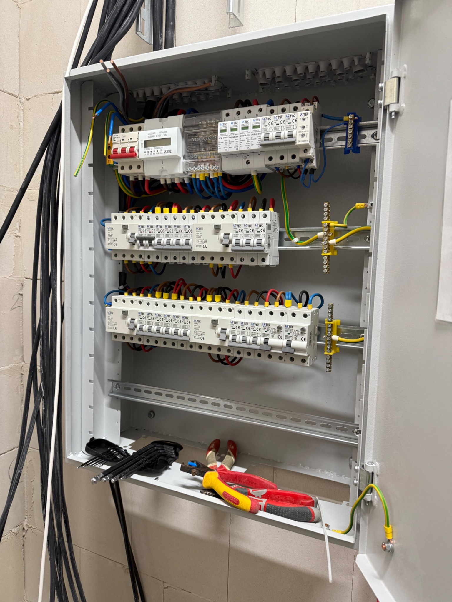 Rozdzielnica elektryczna z zamontowanymi bezpiecznikami, przewodami i licznikiem. Widoczne narzędzia. Instalacja w szarej obudowie na tle ściany z betonu komórkowego.