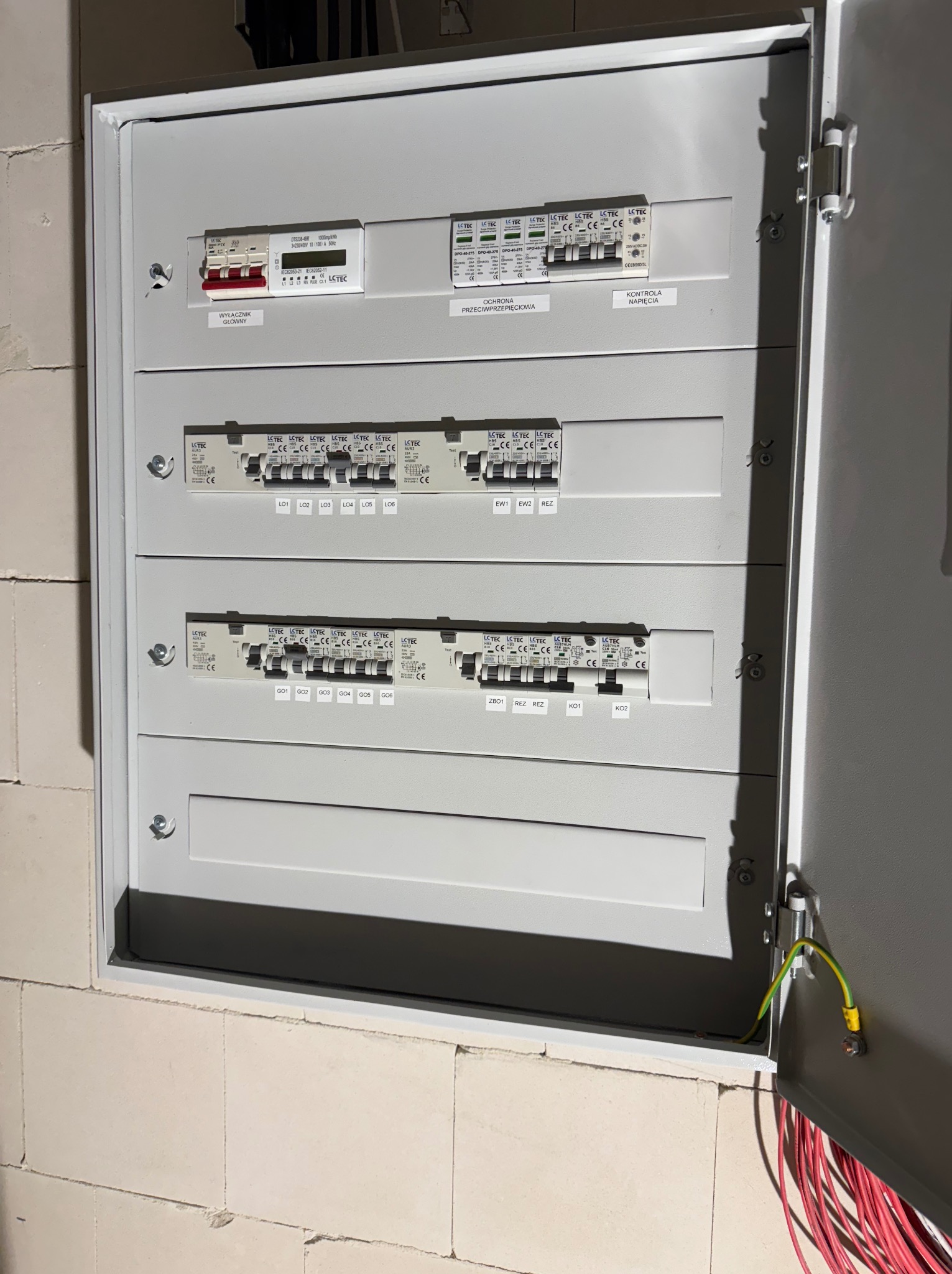 Szara, otwarta skrzynka elektryczna z bezpiecznikami i oznaczeniami, zamontowana na murze z białych bloczków. Widoczne kable wewnątrz.