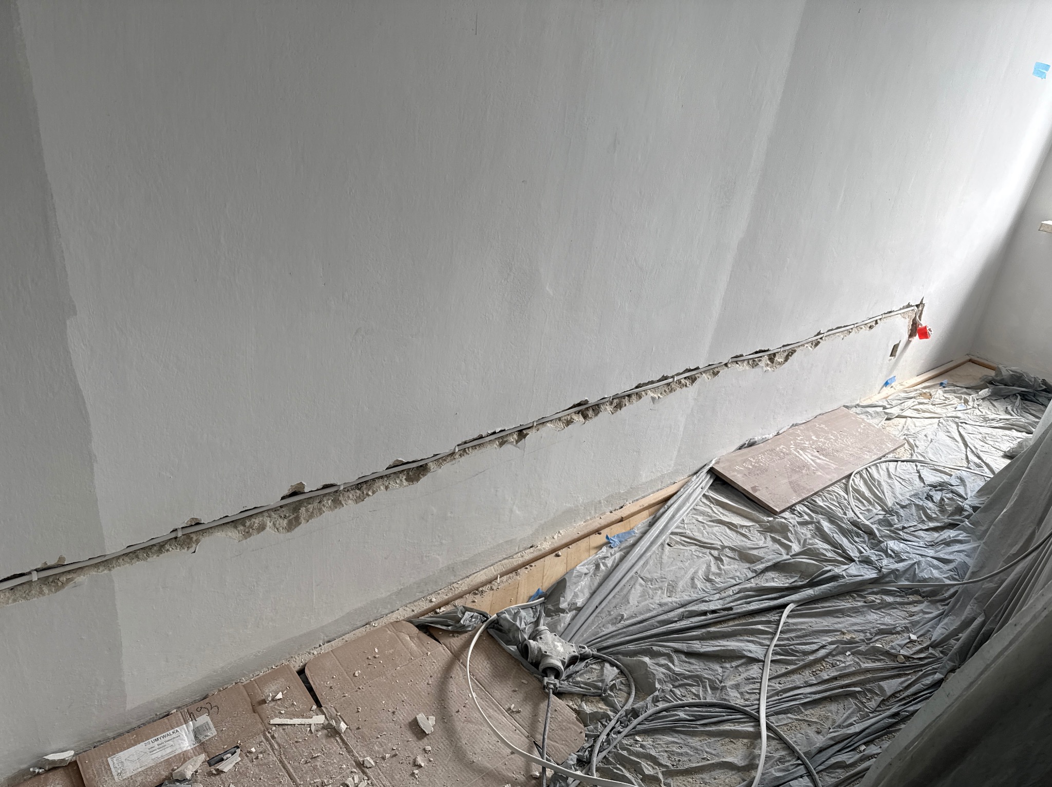 Bruzdowanie ściany pod instalację elektryczną w trakcie remontu. Widoczne kable w korytach, zabezpieczona folią podłoga i fragment ściany z puszką elektryczną.