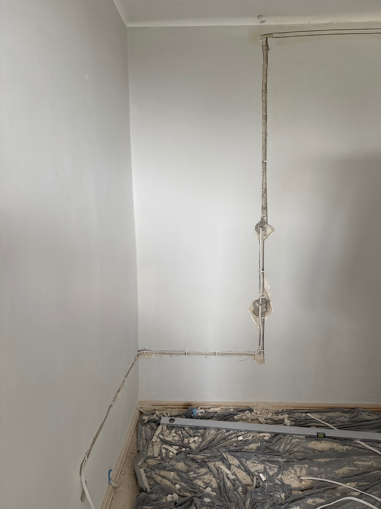 Ściana z bruzdami pod instalację elektryczną, widoczne przewody i puszki. Podłoga zabezpieczona folią, leży poziomica. Etap prac remontowych.