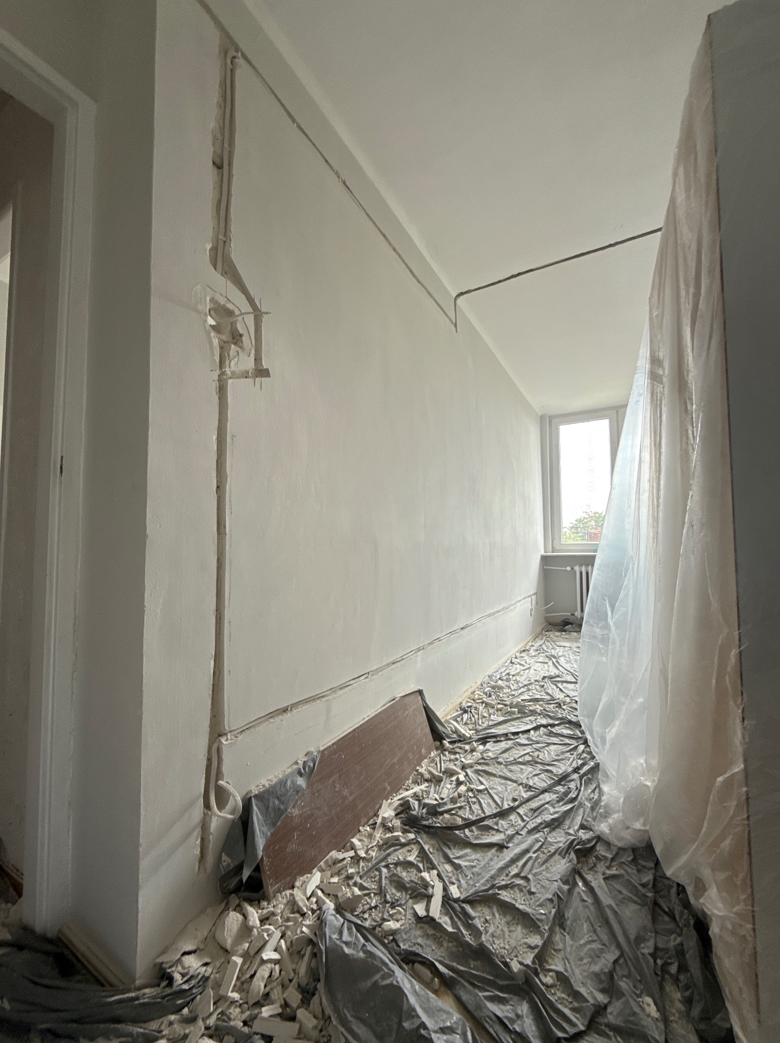 Korytarz w trakcie remontu: ściany z bruzdami pod instalacje elektryczne, podłoga zabezpieczona folią i gruzem, widoczne okablowanie. Etap prac przygotowawczych.