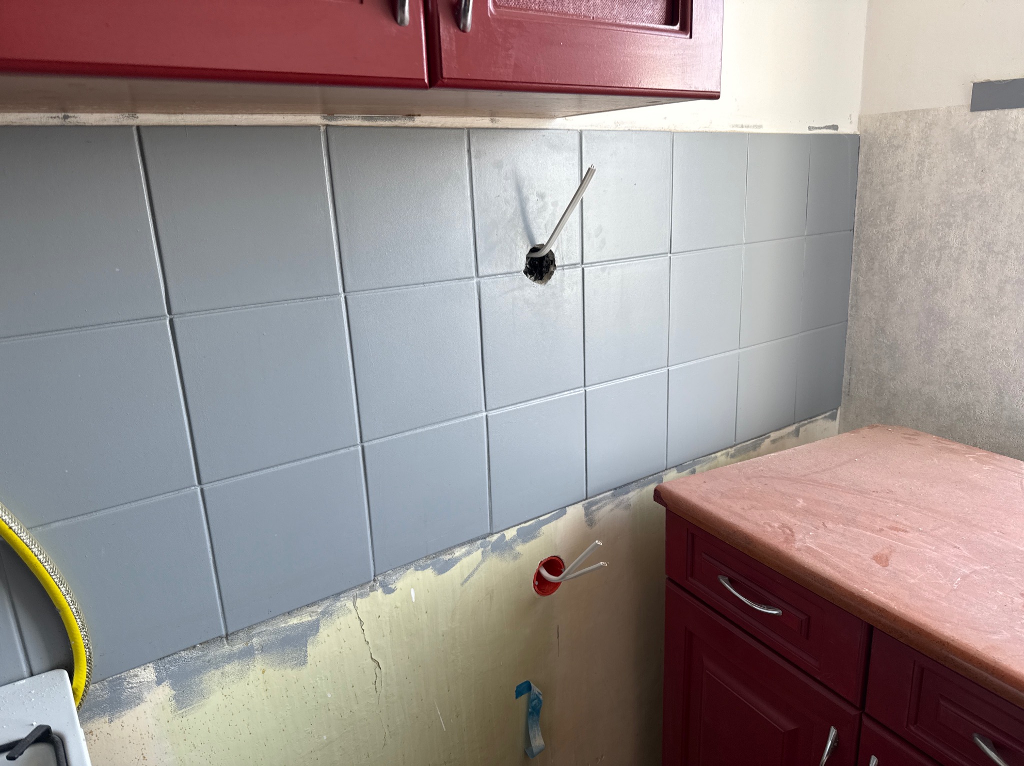 Szare płytki w kuchni, częściowo ułożone, z wystającymi przewodami elektrycznymi. Widoczne czerwone szafki i przygotowane ściany do dalszych prac wykończeniowych.