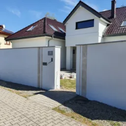 Biały, nowoczesny dom z ciemnobrązowym dachem, widoczny za świeżo pomalowanym, jasnym murem z numerem 10. Puszki z farbą w tle sugerują prace wykończeniowe.