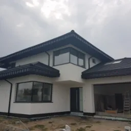 Nowoczesny dom jednorodzinny z białą elewacją i ciemną stolarką okienną. Widoczne detale wykończenia, boczny widok budynku, garaż otwarty, drabina w garażu.