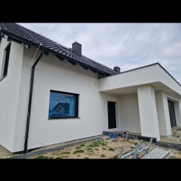 ZK HOUSE ANNA OTOLIŃSKA - Nowoczesny dom z białą elewacją i czarnym dachem. Okno zabezpieczone folią. Widoczne elementy wykończeniowe wokół budynku. Ujęcie z niskiej perspektywy.