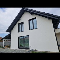 ZK HOUSE ANNA OTOLIŃSKA - Biała elewacja domu jednorodzinnego z czarną stolarką okienną i dachem. Okno na parterze zabezpieczone folią malarską. Widoczne wykończenie terenu wokół budynku.