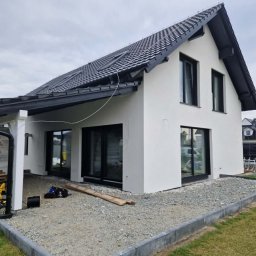 ZK HOUSE ANNA OTOLIŃSKA - Nowoczesny dom jednorodzinny z białą elewacją i czarną stolarką okienną. Widoczne narzędzia sugerują prace wykończeniowe.