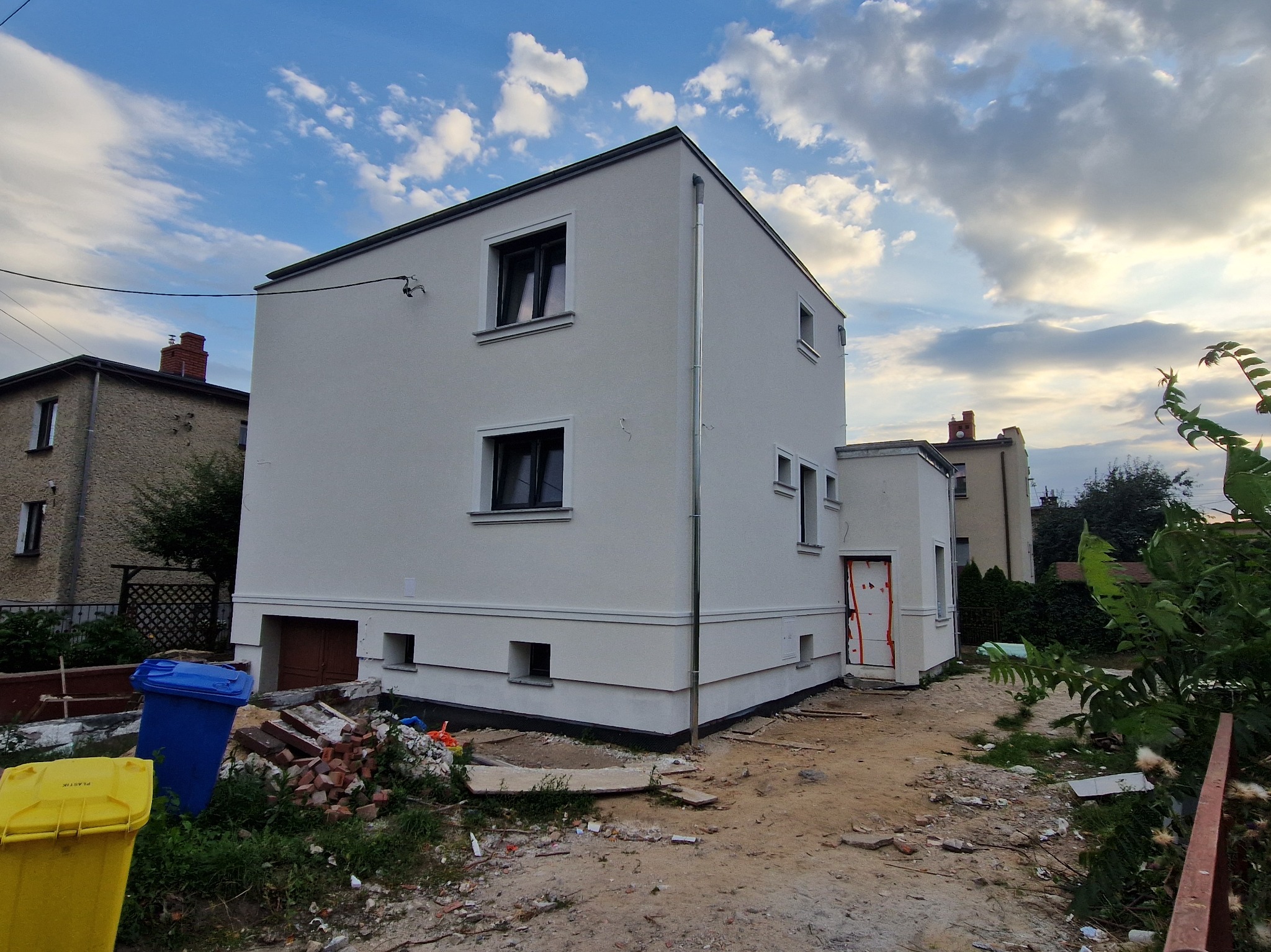 Nowoczesny, dwukondygnacyjny budynek po renowacji elewacji, z widocznymi oknami i świeżo pomalowaną fasadą. W tle błękitne niebo z chmurami. Na pierwszym planie gruz i śmietniki.