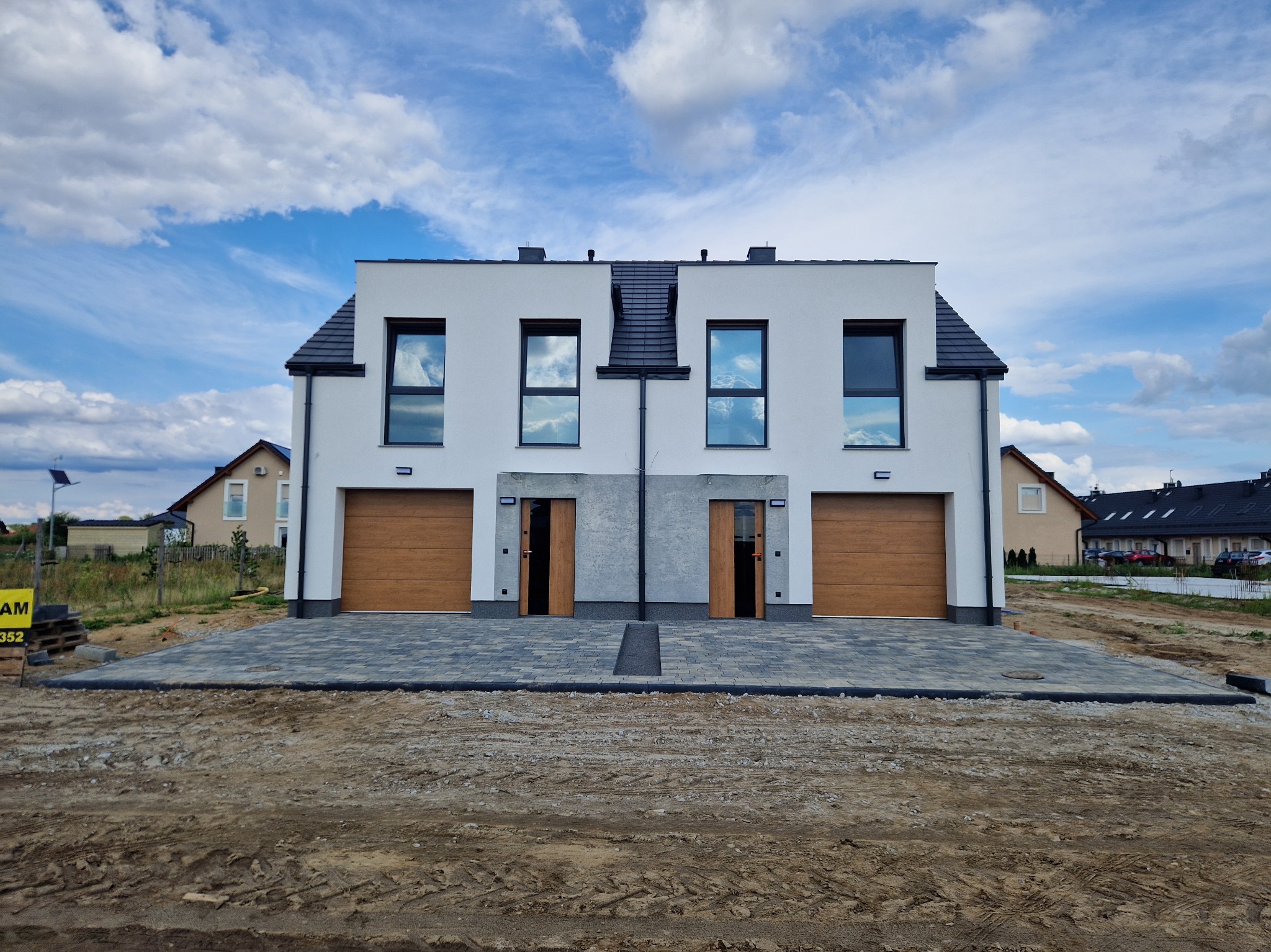 Bliźniak z białą elewacją i drewnianymi bramami garażowymi, widziany z poziomu gruntu na tle błękitnego nieba z chmurami. Ułożona kostka brukowa przed budynkiem.