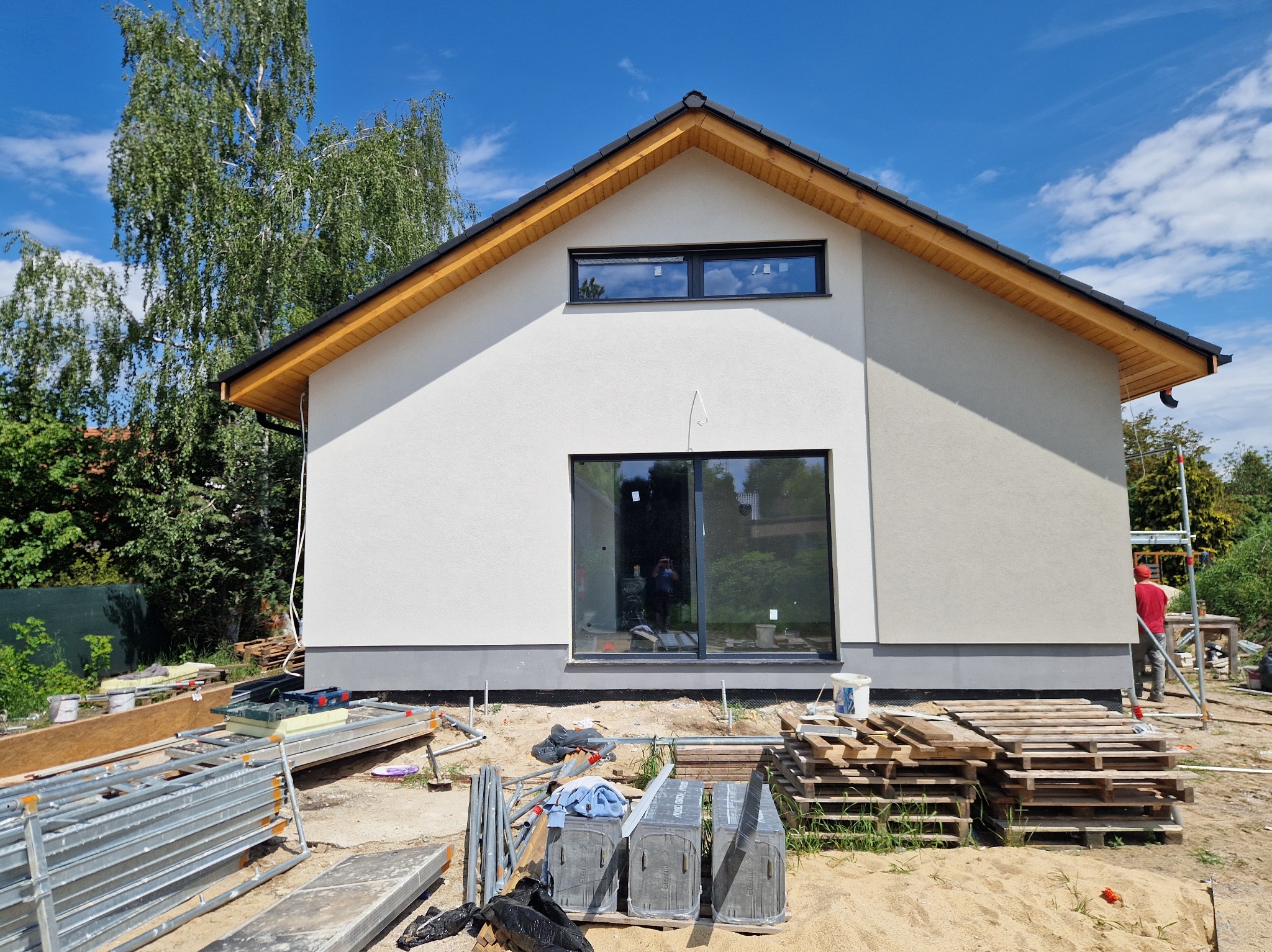 Nowoczesny dom jednorodzinny z jasną elewacją i ciemną stolarką okienną. Widoczne rusztowania i materiały budowlane sugerują prace wykończeniowe.