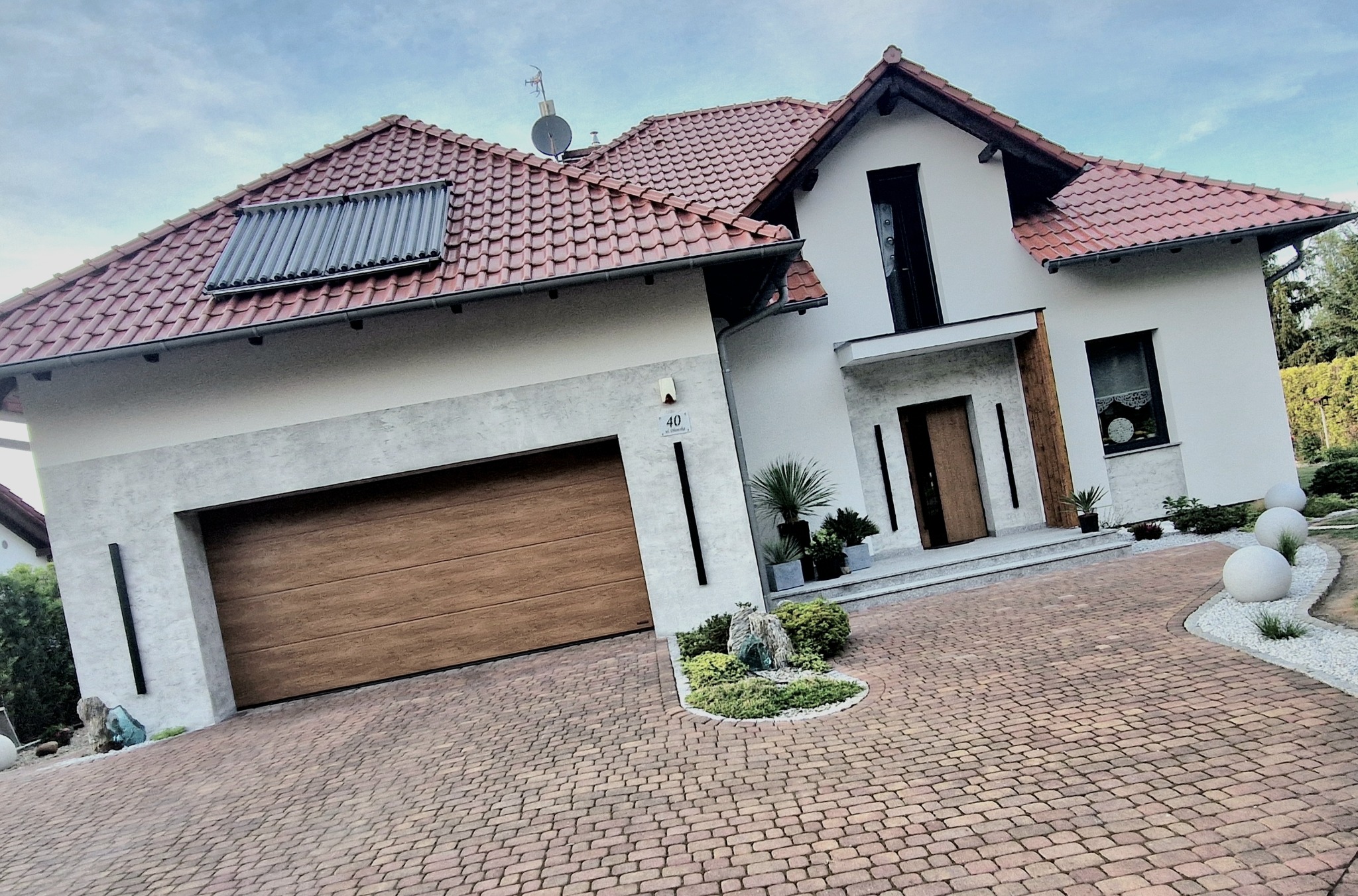 Nowoczesny dom jednorodzinny z brązową bramą garażową, elewacją w odcieniach bieli i beżu oraz dachem z czerwonej dachówki. Widoczne panele słoneczne i ozdobne elementy.