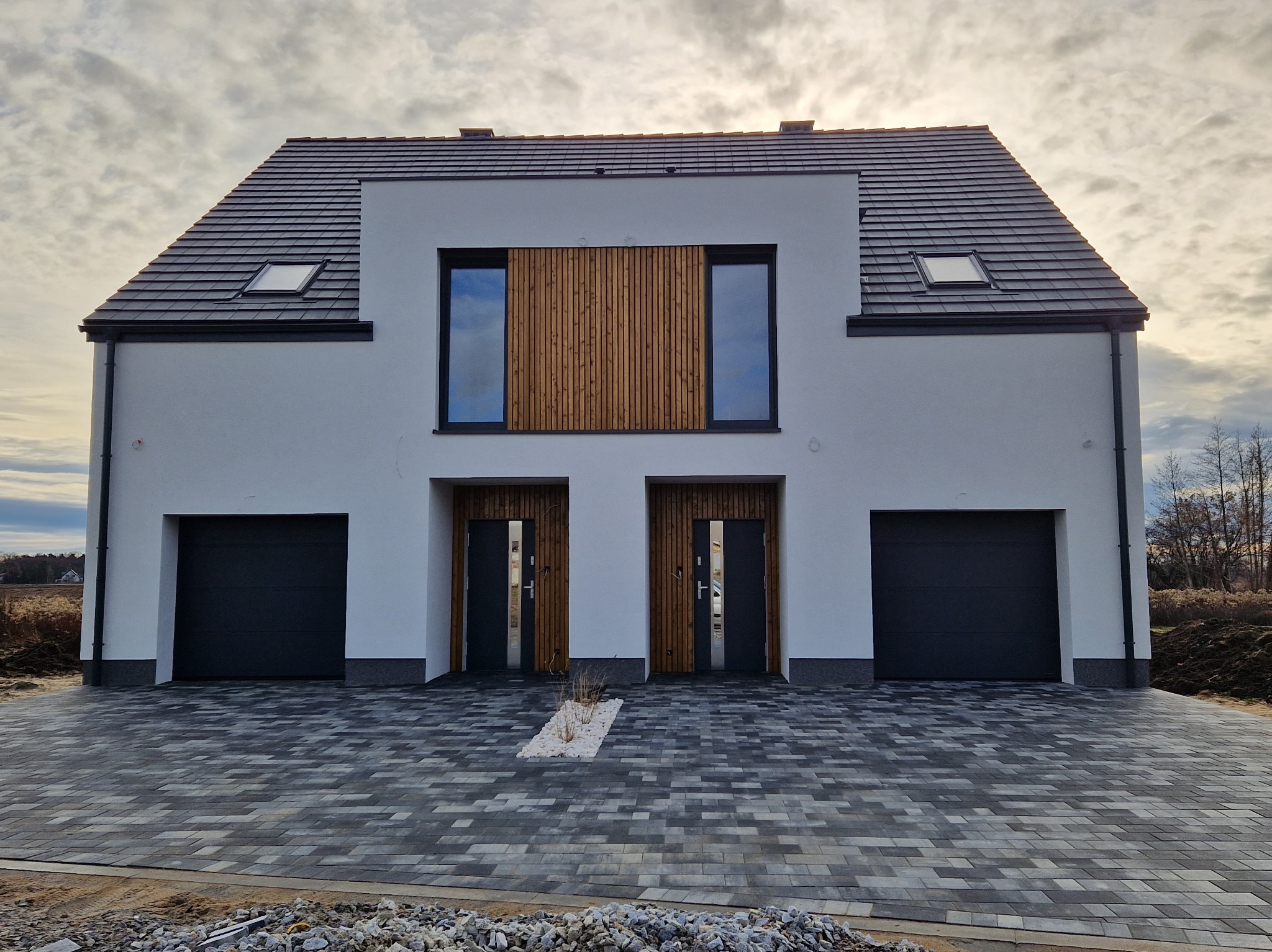 Nowoczesny dom jednorodzinny z szarą dachówką i elementami drewnianymi na elewacji, widok z zewnątrz na dwuskrzydłowe drzwi wejściowe i podjazd z kostki brukowej.