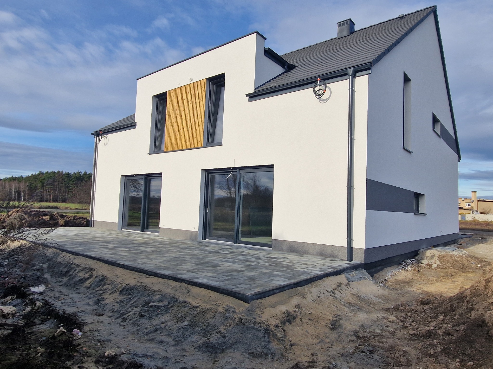 Nowoczesny dom jednorodzinny z białymi ścianami i szarą kostką brukową na tarasie. Na elewacji widoczne drewniane żaluzje okienne i szare akcenty.