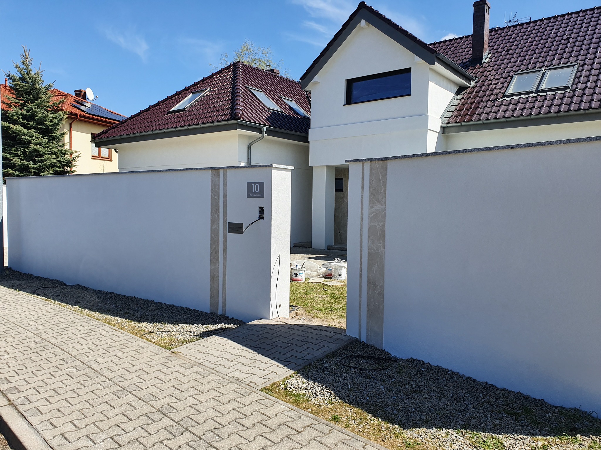 Biały, nowoczesny dom z ciemnobrązowym dachem, widoczny za świeżo pomalowanym, jasnym murem z numerem 10. Puszki z farbą w tle sugerują prace wykończeniowe.