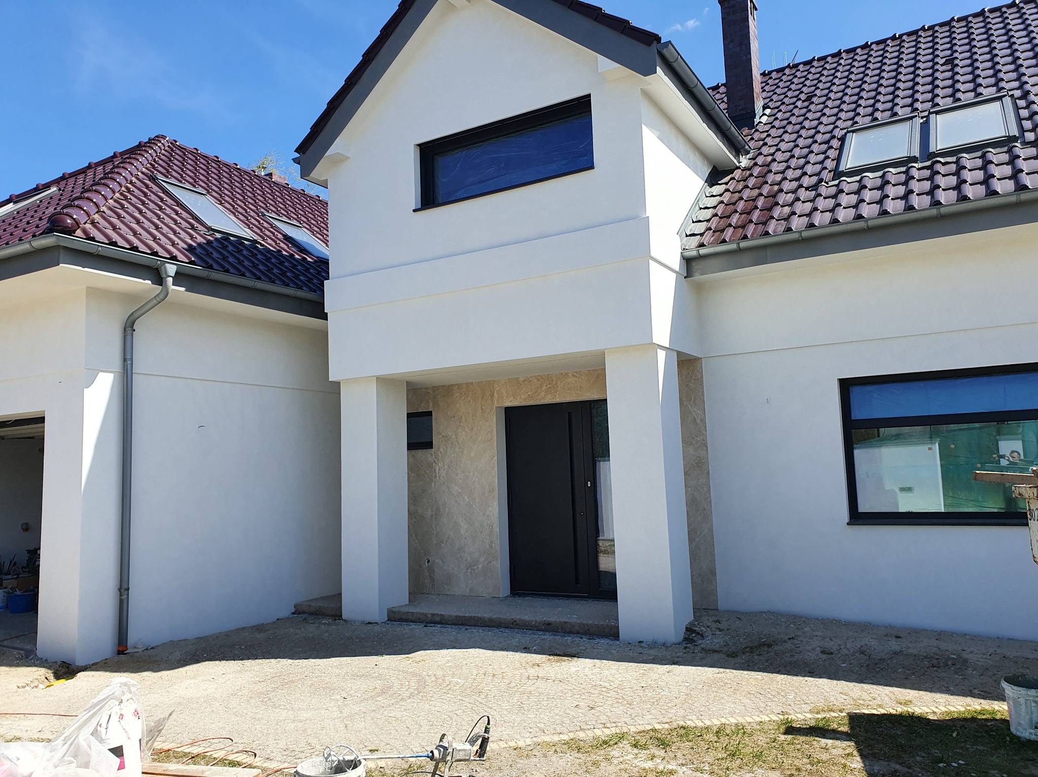 Biały dom jednorodzinny z ciemną stolarką okienną i drzwiami wejściowymi, widok na elewację po renowacji, sprzęt malarski na pierwszym planie.