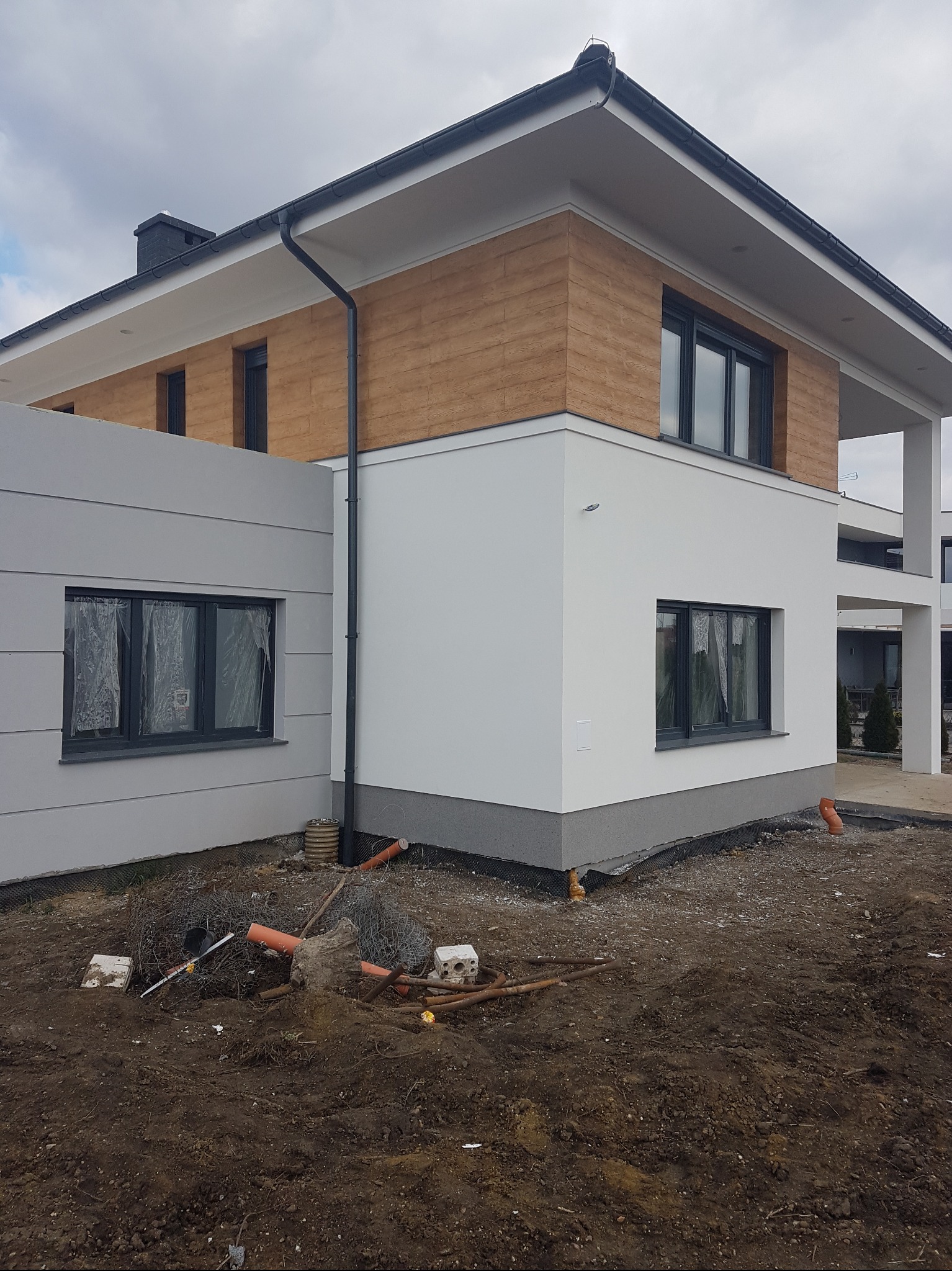 Nowoczesny dom z elewacją w odcieniach bieli i drewna, czarnymi oknami, widoczne prace ziemne wokół budynku. Górna część elewacji wykończona imitacją drewna.