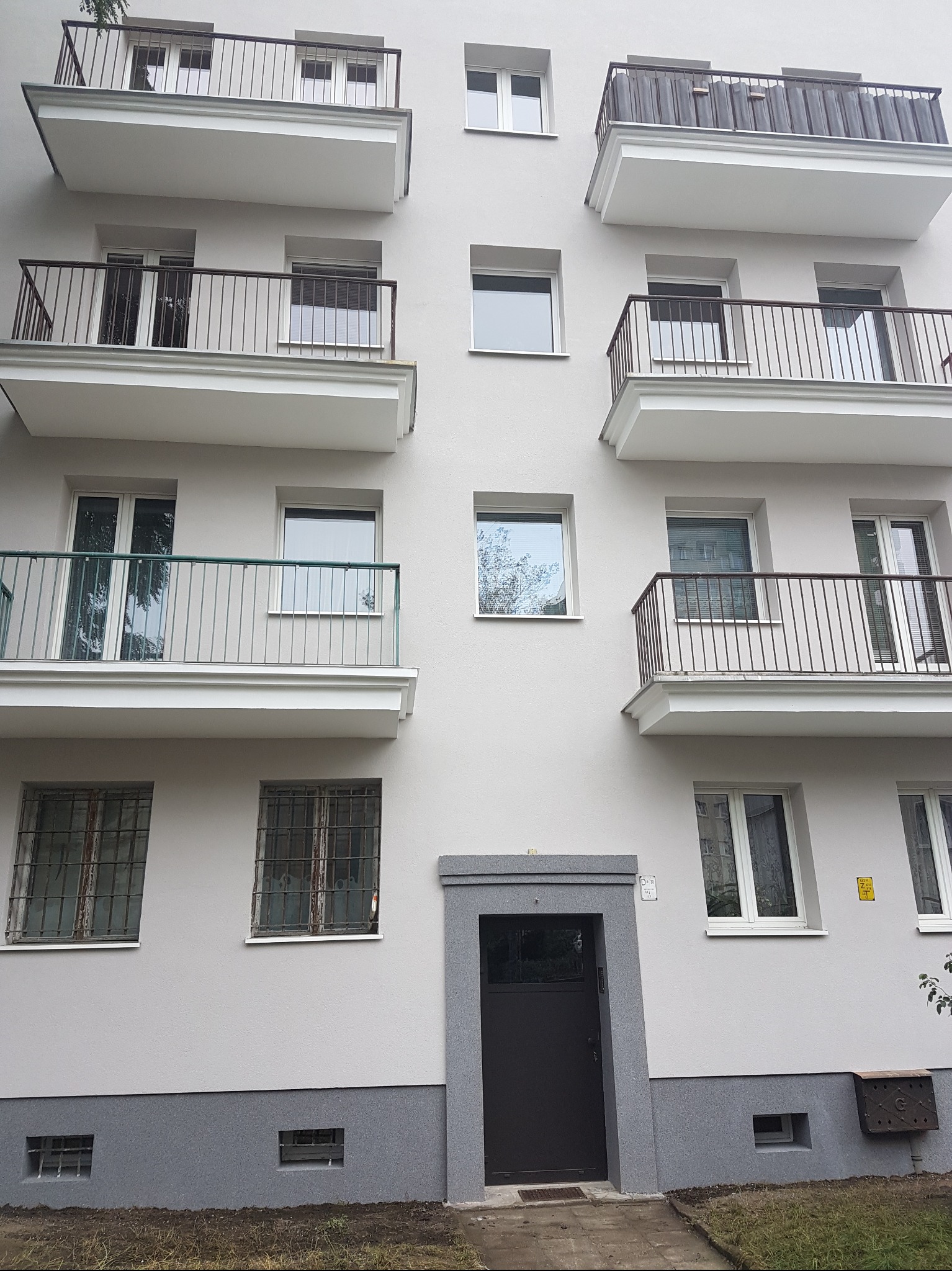 Elewacja budynku po renowacji z balkonami o różnych balustradach i odnowioną strefą wejścia z ciemnymi drzwiami i szarą lamperią.