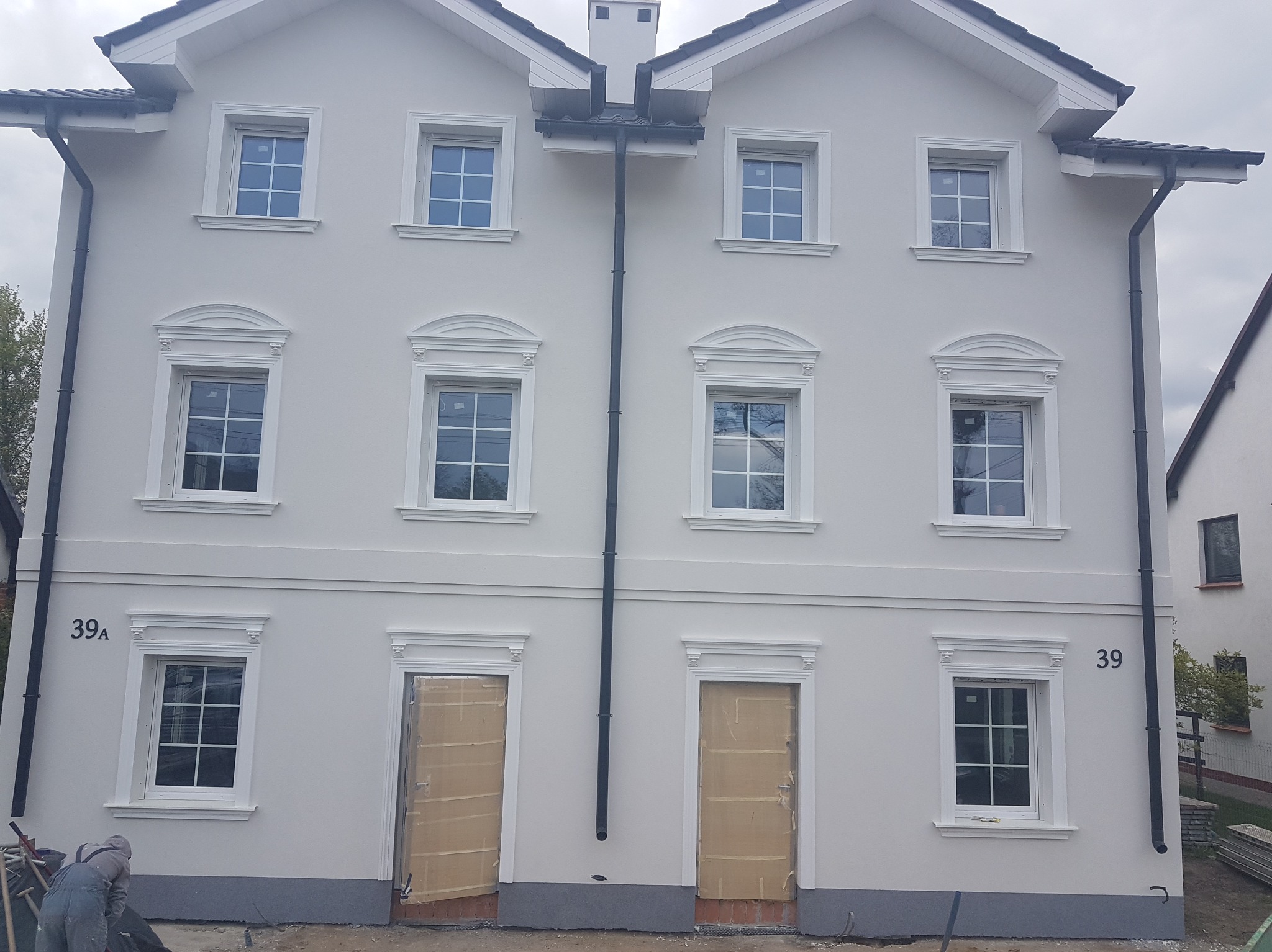 Odnowiona elewacja bliźniaka z dekoracyjnymi obramowaniami okien i drzwi, świeżo pomalowana na jasny kolor. Widoczne numery budynków 39 i 39A.