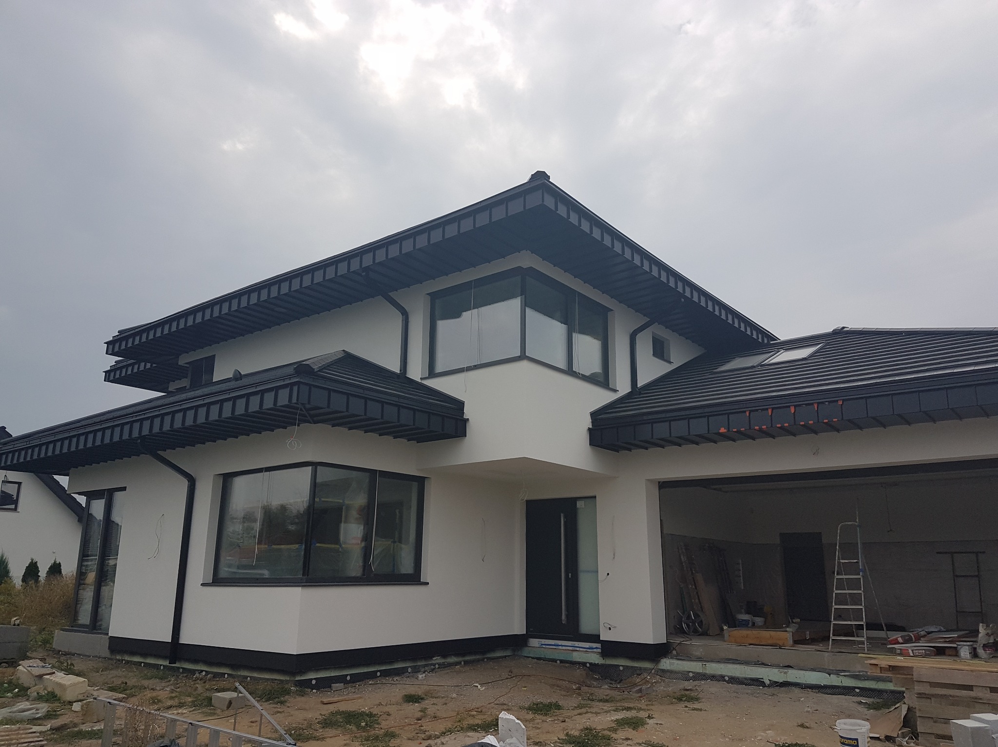 Nowoczesny dom jednorodzinny z białą elewacją i ciemną stolarką okienną. Widoczne detale wykończenia, boczny widok budynku, garaż otwarty, drabina w garażu.