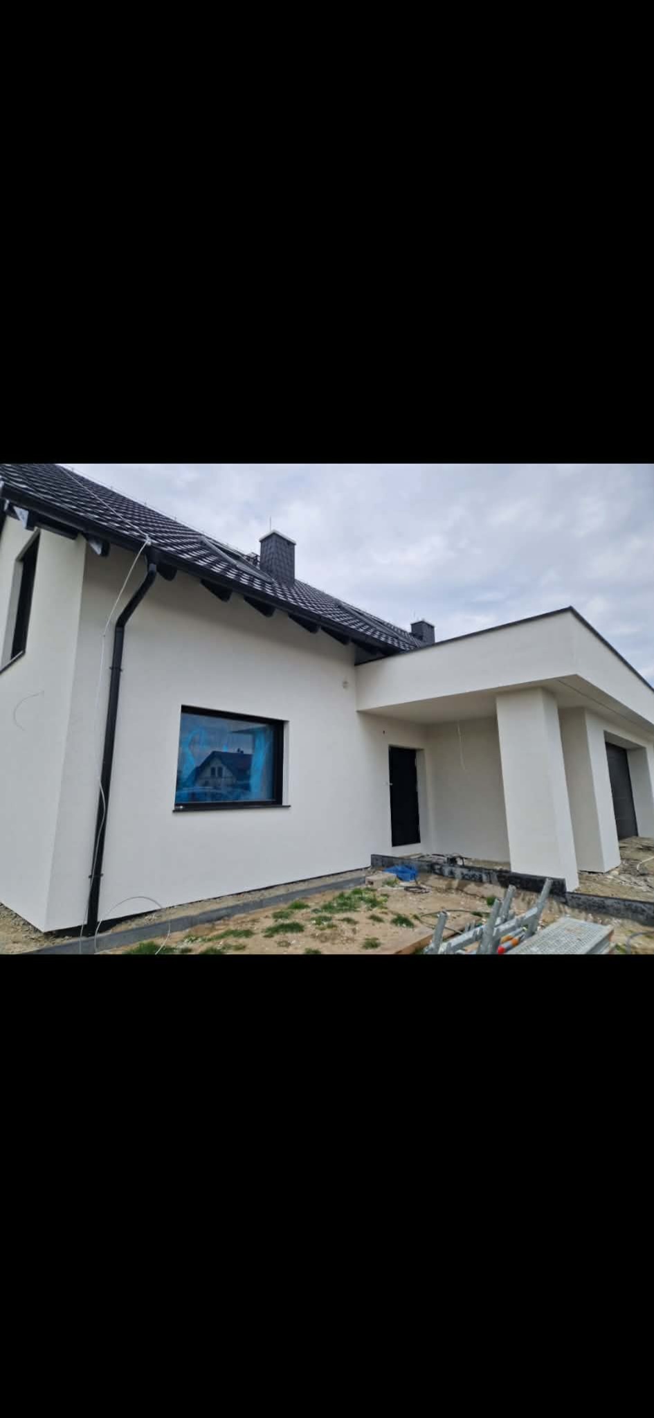 Nowoczesny dom z białą elewacją i czarnym dachem. Okno zabezpieczone folią. Widoczne elementy wykończeniowe wokół budynku. Ujęcie z niskiej perspektywy.