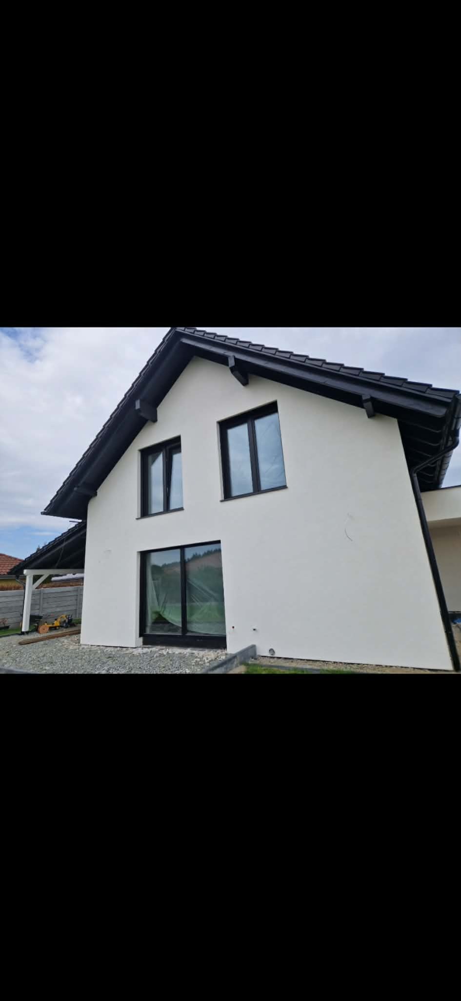 Biała elewacja domu jednorodzinnego z czarną stolarką okienną i dachem. Okno na parterze zabezpieczone folią malarską. Widoczne wykończenie terenu wokół budynku.