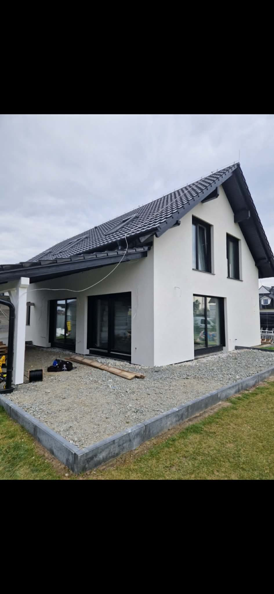 Nowoczesny dom jednorodzinny z białą elewacją i czarną stolarką okienną. Widoczne narzędzia sugerują prace wykończeniowe.