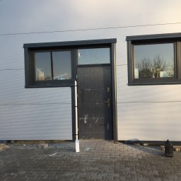 Usługi ogólnobudowlane Kamil Hintzke - Elewacja budynku przemysłowego z szarej blachy trapezowej, z dwoma oknami z czarnymi ramami i wejściem z czarnymi drzwiami, widok na kostkę brukową.