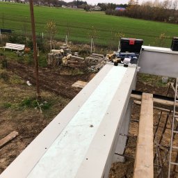 Usługi ogólnobudowlane Kamil Hintzke - Widok z góry na konstrukcję betonową z izolacją termiczną w trakcie budowy, narzędzia i skrzynki narzędziowe na górze, drabina oparta o konstrukcję, w tle zielone pole i zabudowania.