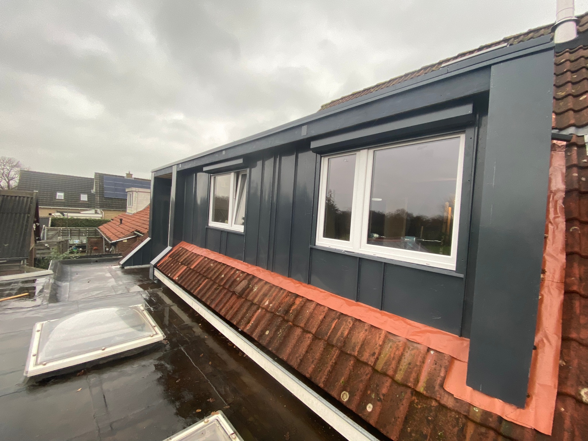 Szare obicie domu z białymi oknami, widok z dachu z lukarną i starymi dachówkami, pochmurny dzień.