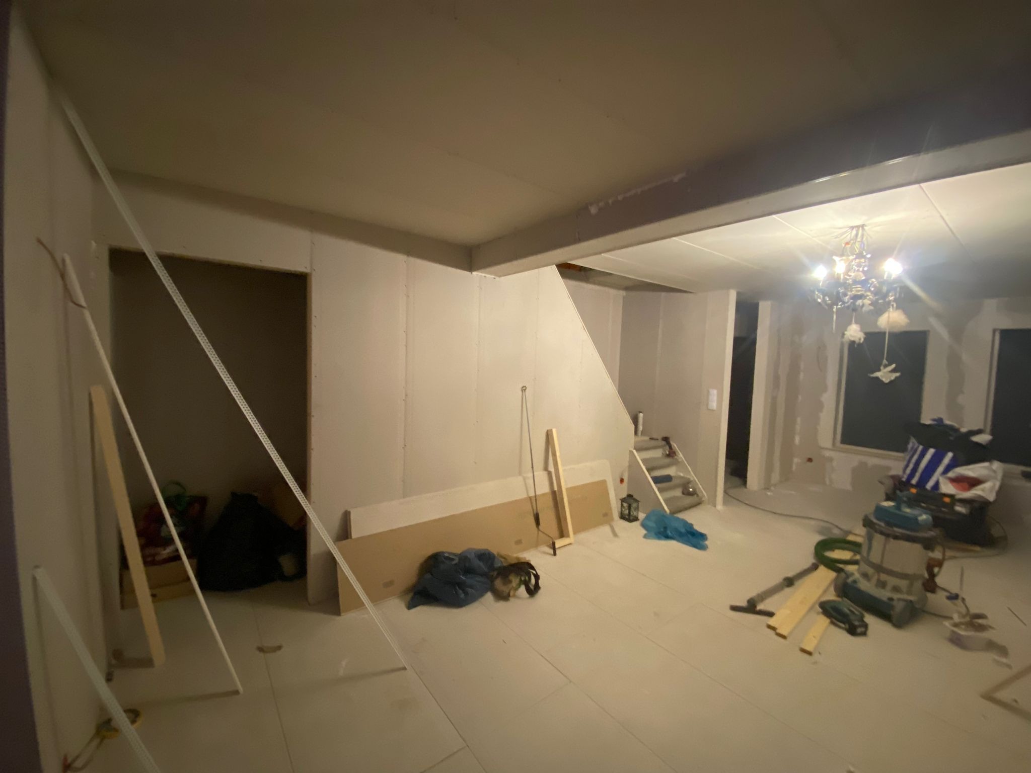 Wnętrze w trakcie remontu: ściany obłożone płytami gipsowo-kartonowymi, widoczne profile, narzędzia i odkurzacz przemysłowy, surowe wykończenie podłogi, w tle schody i okno.