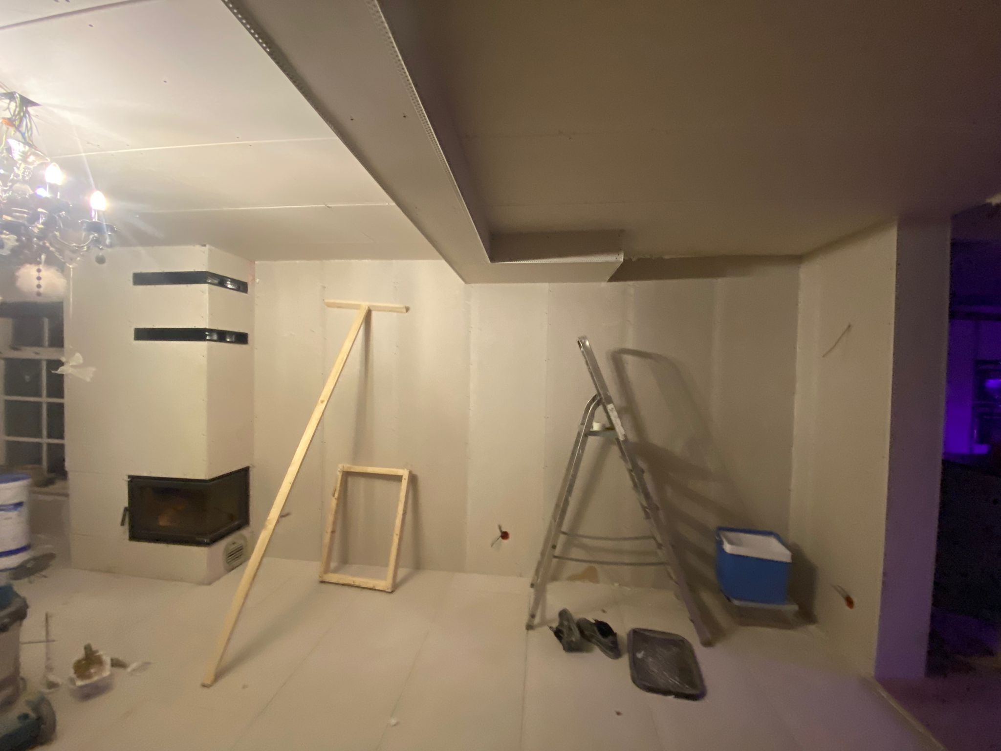 Wnętrze w trakcie prac wykończeniowych: ściany obłożone płytami gipsowo-kartonowymi, widoczny kominek z czarnymi detalami, drabina, drewniane stelaże, narzędzia i wiadro z farbą. Sufit częściowo...