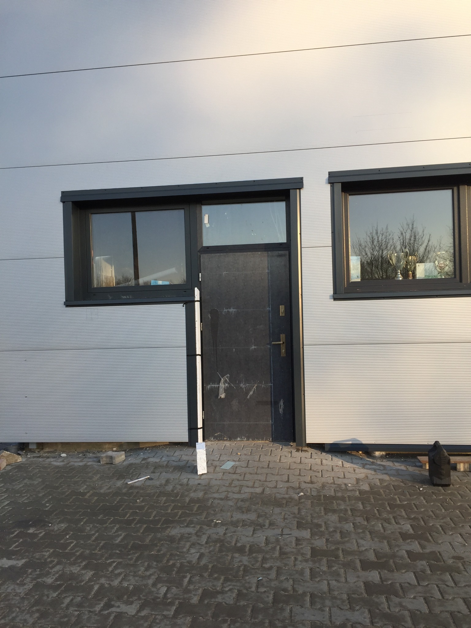 Elewacja budynku przemysłowego z szarej blachy trapezowej, z dwoma oknami z czarnymi ramami i wejściem z czarnymi drzwiami, widok na kostkę brukową.
