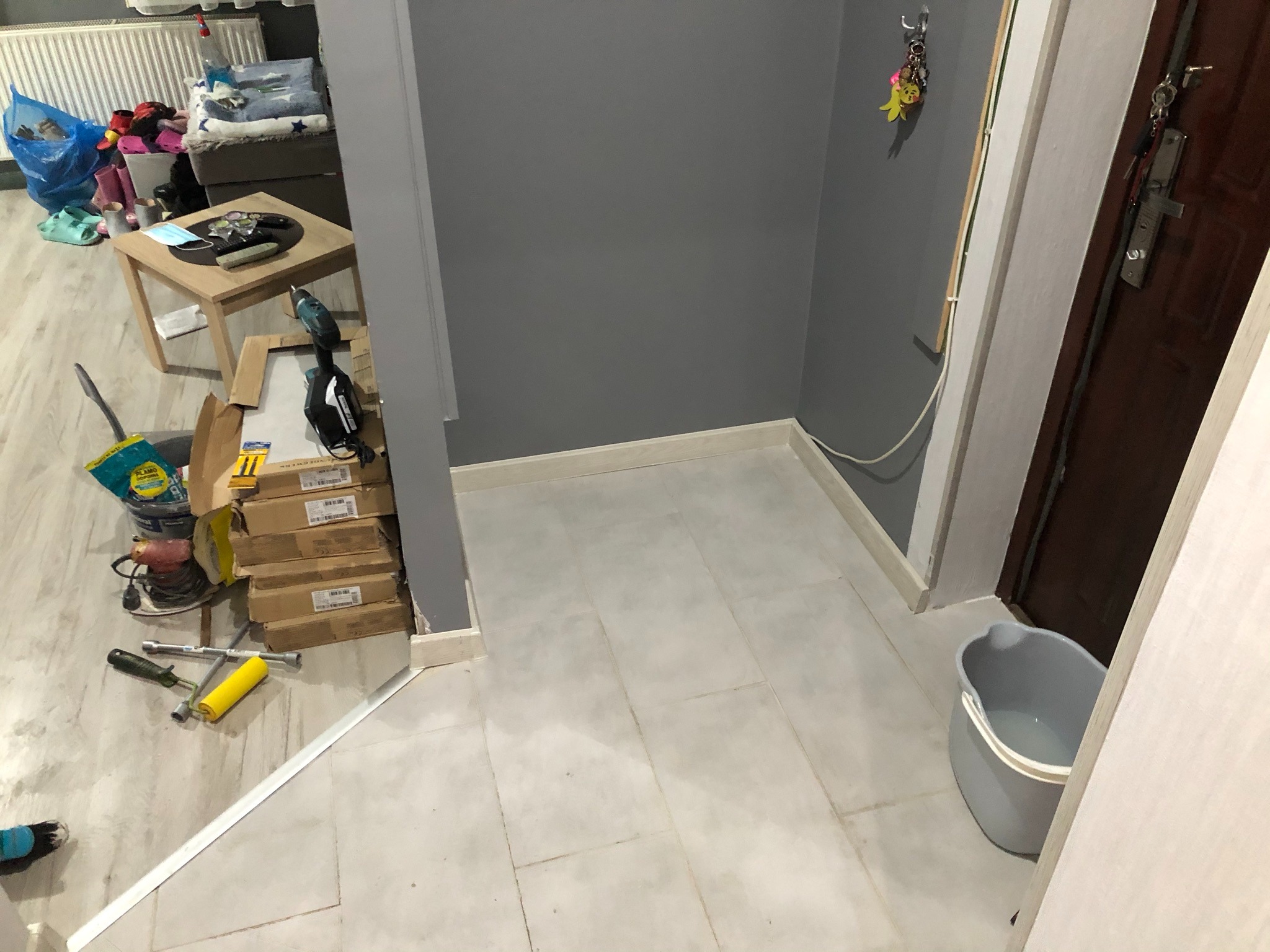 Przygotowanie do malowania ścian w pomieszczeniu: widoczne narzędzia malarskie, pędzle, wałki, farby w wiadrach i kartony, szare ściany i jasne płytki podłogowe.