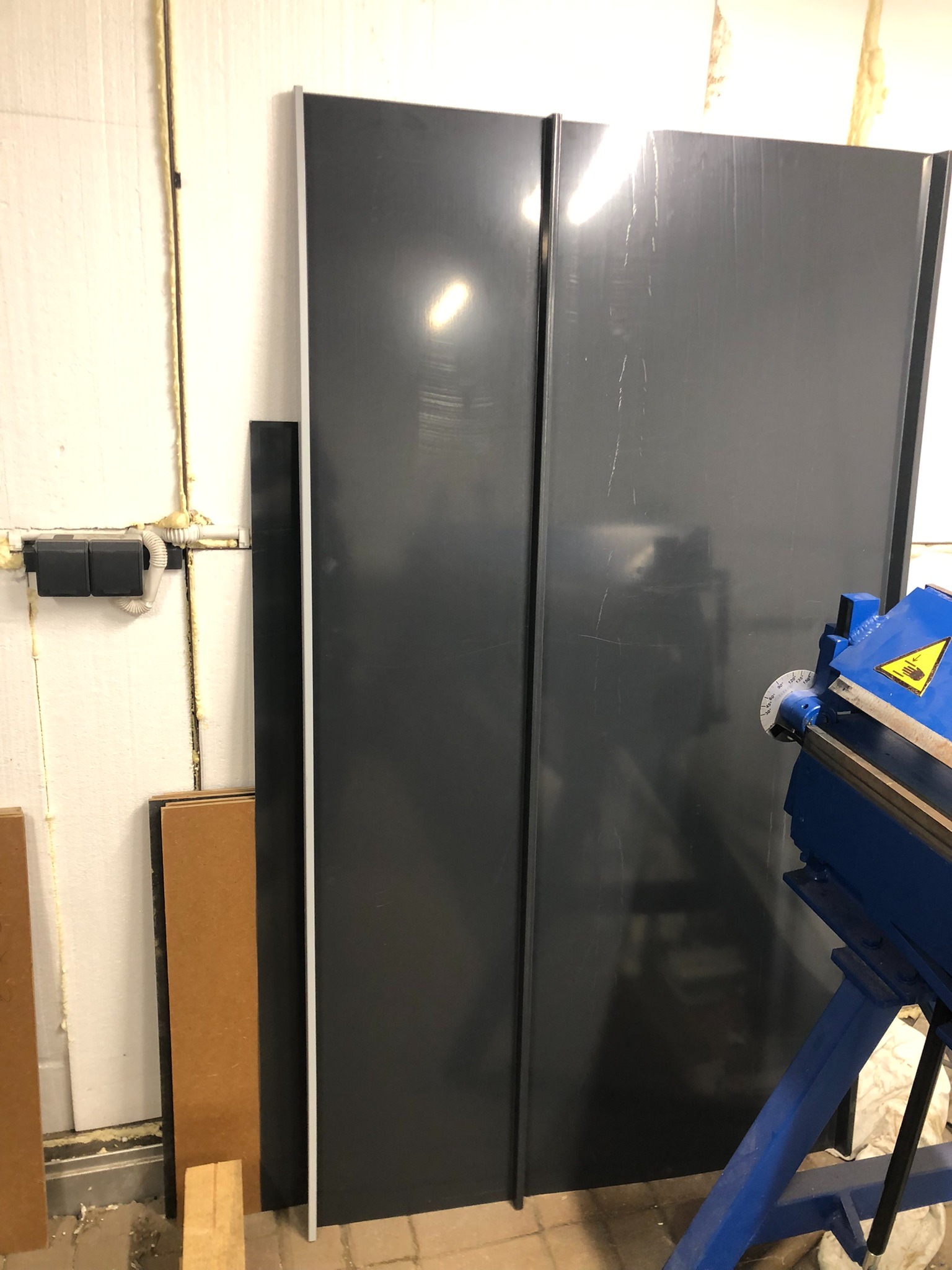 Dwa ciemne panele blaszane stojące przy ścianie, obok niebieska giętarka do blachy, widoczne ślady użytkowania na panelach.