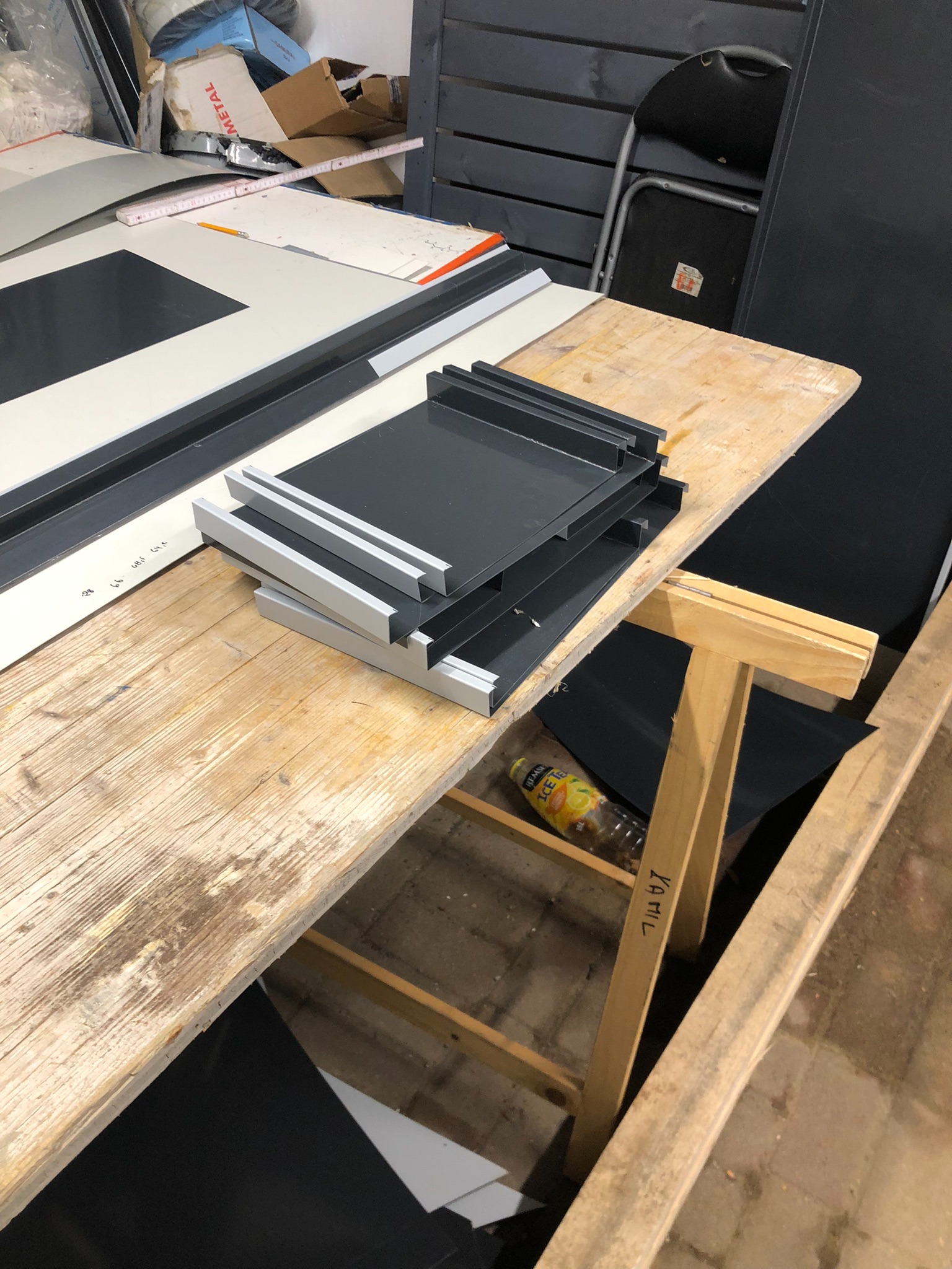 Stos szarych, metalowych rynien na drewnianym stole warsztatowym, obok arkuszy blachy i butelki z napojem.