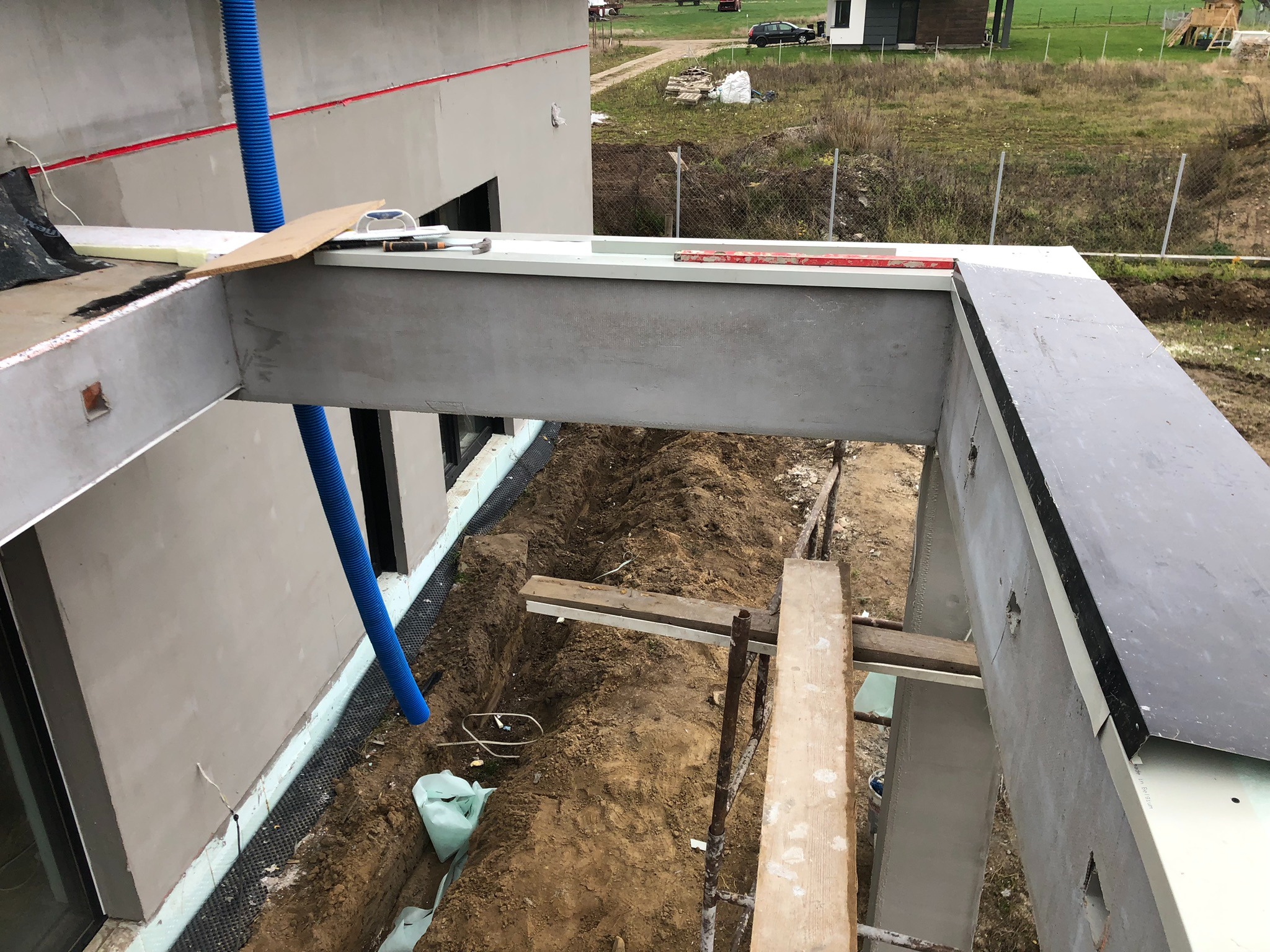 Surowy stan elewacji budynku w trakcie prac wykończeniowych, widoczne ocieplenie fundamentów, szara elewacja, niebieska rura wentylacyjna i wykop w ziemi.